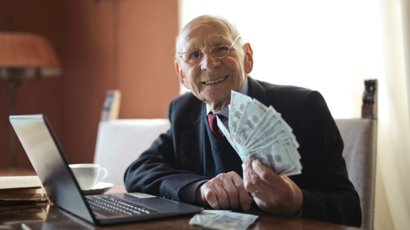 happy senior businessman holding money in hand while working on laptop at table
